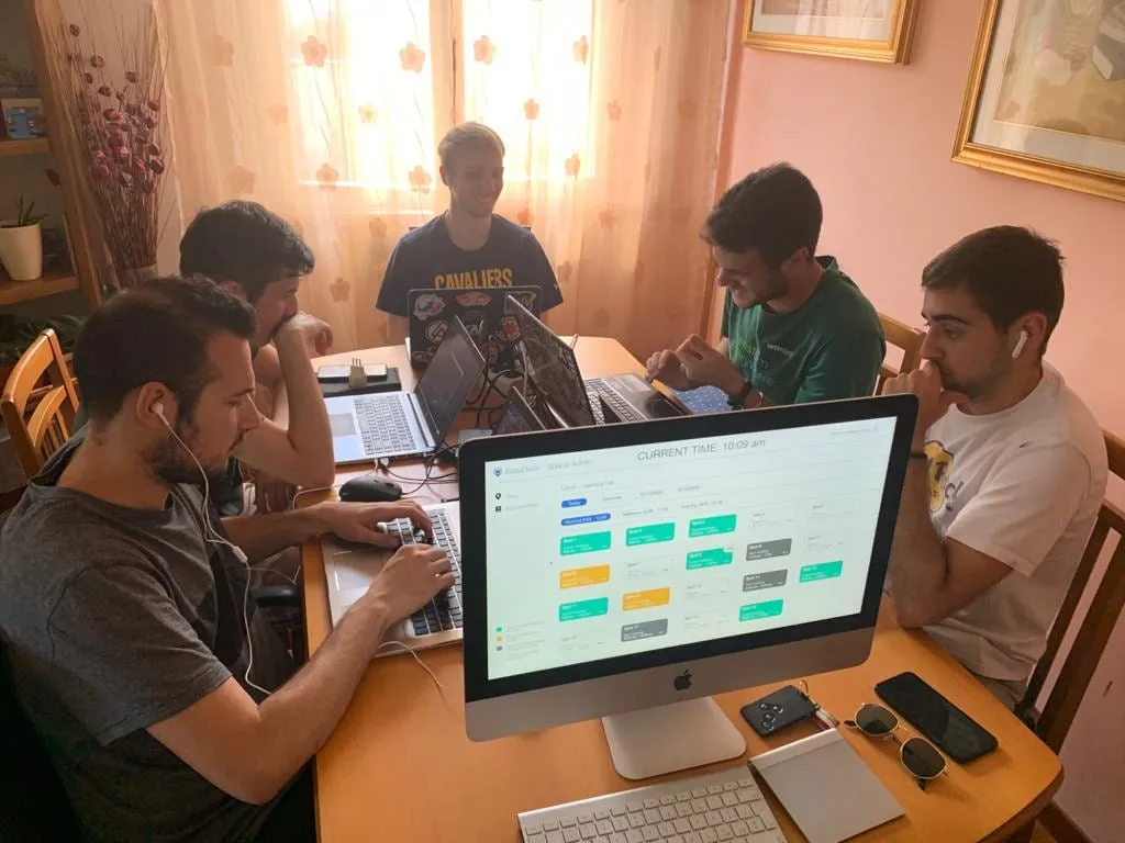 Five of the team around a dining table with laptops, an iMac in front showing the EasyDesk admin grid of bookable spots