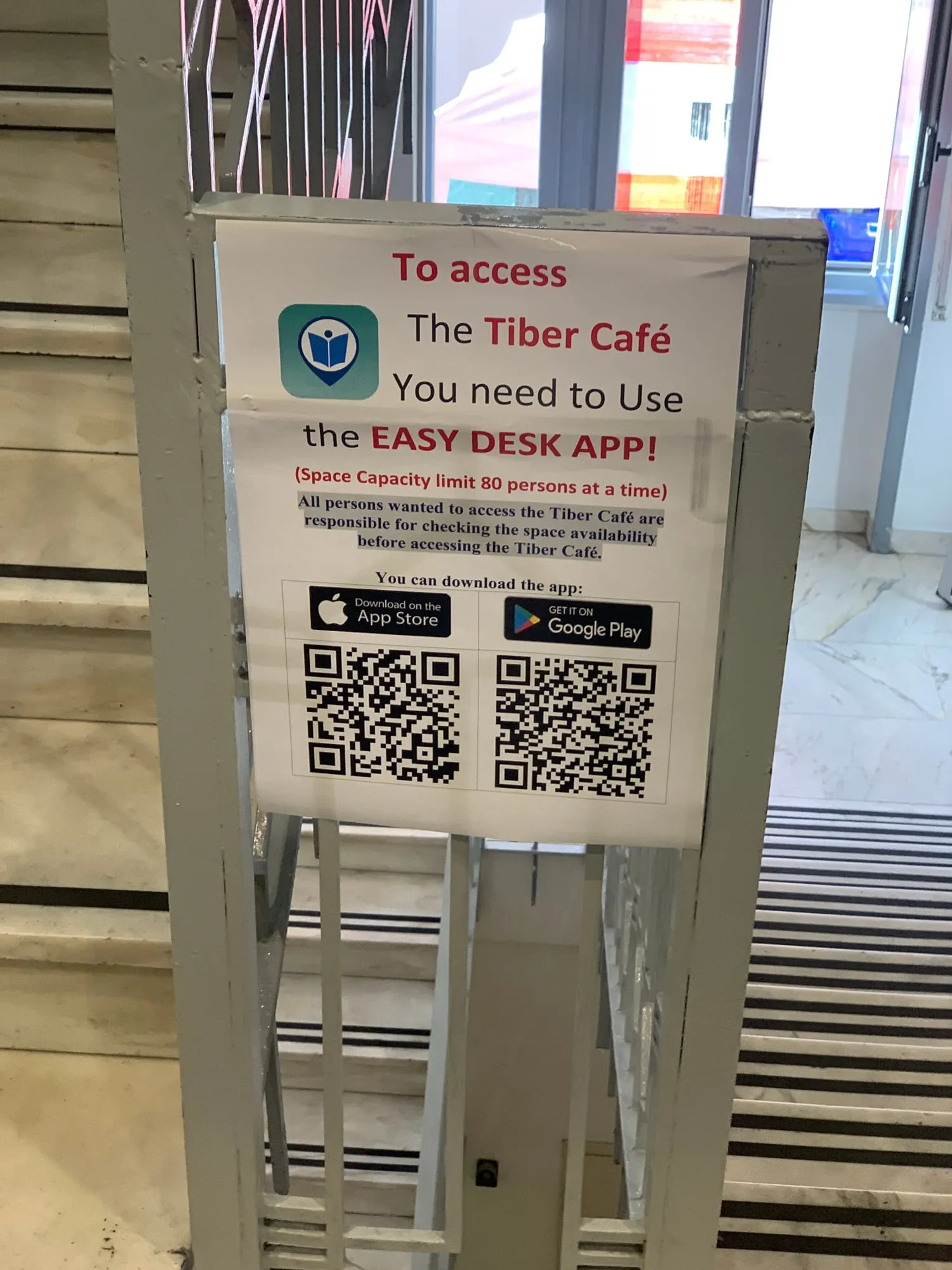 A printed sign on a staircase railing: 'To access the Tiber Café you need to use the EasyDesk app', with a capacity limit of 80 people and download QR codes